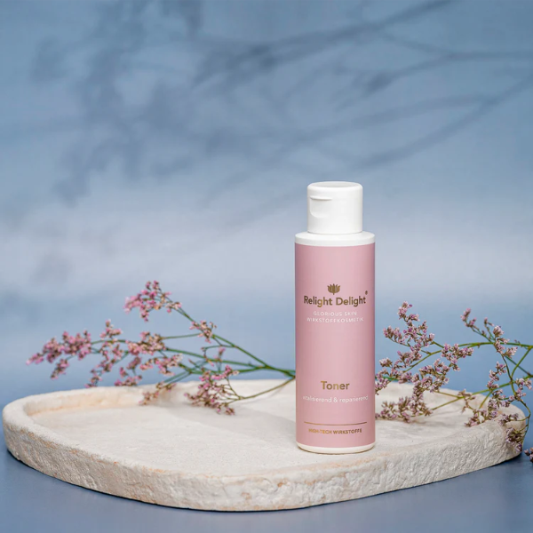 Tube in edlem Rosé mit goldener Schrift: Glorious Skin - Toner - unbeduftet - von Relight Delight steht auf einem flachen Stein. Daneben Blüten und der Hintergrund zart in blau