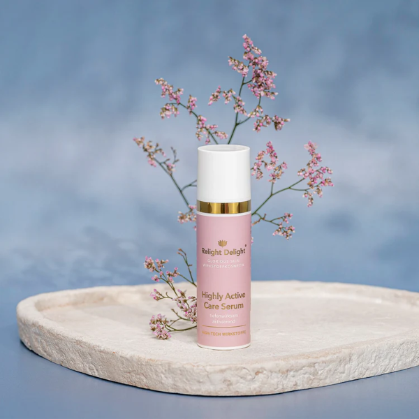 Edle Tube in Rosé mit goldener und weißer Aufschrift: Glorious Skin Highly Active Care Serum, tiefenwirksam und aktivierend High Tech Wirkstoffkosmetik von  Relight Delight auf hellem, flachen Stein stehend mit zarten rosa Blüten und hellblauem Hintergrund