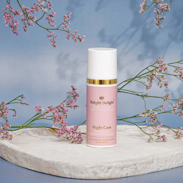 Edle Tube in Rosé mit goldener und weißer Aufschrift: Glorious Skin - Night Care , tiefenwirksam regenerierend und schützend 50ml, auf basis kaltgepresster Bio Wirköle von - Relight Delight auf hellem flachen Stein stehend mit zarten rosafarbenen Blüten.
