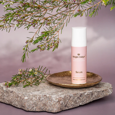 
Auf dem Bild ist eine Flasche Relight Delight Serum zu sehen, die auf einer kleinen, goldenen Schale steht. Diese Schale befindet sich wiederum auf einem groben, grauen Steinblock. Das Serum-Fläschchen ist schlicht und modern gestaltet: Es hat einen weißen Deckel und einen rosafarbenen Körper mit goldenem Aufdruck. Um das Produkt herum sind Zweige mit feinen, nadelförmigen Blättern und kleinen, zartrosa Blüten arrangiert, die dem Bild eine natürliche und sanfte Note verleihen. 
