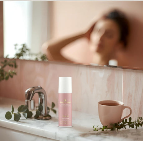 Morgenroutine im Badezimmer darstellt.
Im Vordergrund ist eine weiße Marmorablage zu sehen. Darauf stehen:
Eine rosèfarbene Tube des Beauty Basics Serum  mit weißem Deckel.
Eine rosèfarbene Tasse mit einem Tee.
Zweig mit Eukalyptusblättern, der dekorativ um die Tasse und den Wasserhahn drapiert ist.
Im Hintergrund ist ein großer Spiegel, der einen rosafarbenen Marmorwaschtisch und einen Teil des Gesichts einer Person unscharf reflektiert, die sich die Haare richtet. Die Beleuchtung ist weich und sanft.
