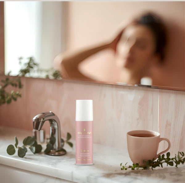 Morgenroutine im Badezimmer darstellt.
Im Vordergrund ist eine weiße Marmorablage zu sehen. Darauf stehen:
Eine rosèfarbene Tube des Beauty Basics Serum  mit weißem Deckel.
Eine rosèfarbene Tasse mit einem Tee.
Zweig mit Eukalyptusblättern, der dekorativ um die Tasse und den Wasserhahn drapiert ist.
Im Hintergrund ist ein großer Spiegel, der einen rosafarbenen Marmorwaschtisch und einen Teil des Gesichts einer Person unscharf reflektiert, die sich die Haare richtet. Die Beleuchtung ist weich und sanft.