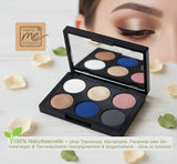 Eine offene schwarze Lidschattenpalette von "That's me organic" mit sechs kreisförmigen Farbtönen: Weiß, glänzendes Gold, mattes Blau, schimmerndes Bronze, Rosé und Anthrazit. Die Palette liegt auf einer hellen Holzoberfläche, umgeben von cremefarbenen Blütenblättern und grünen Blättern. Oben links ist das originale "That's me organic"-Logo. 100% Naturkosmetik – ohne Titandioxid, Mikroplastik, Parabene oder Silikone