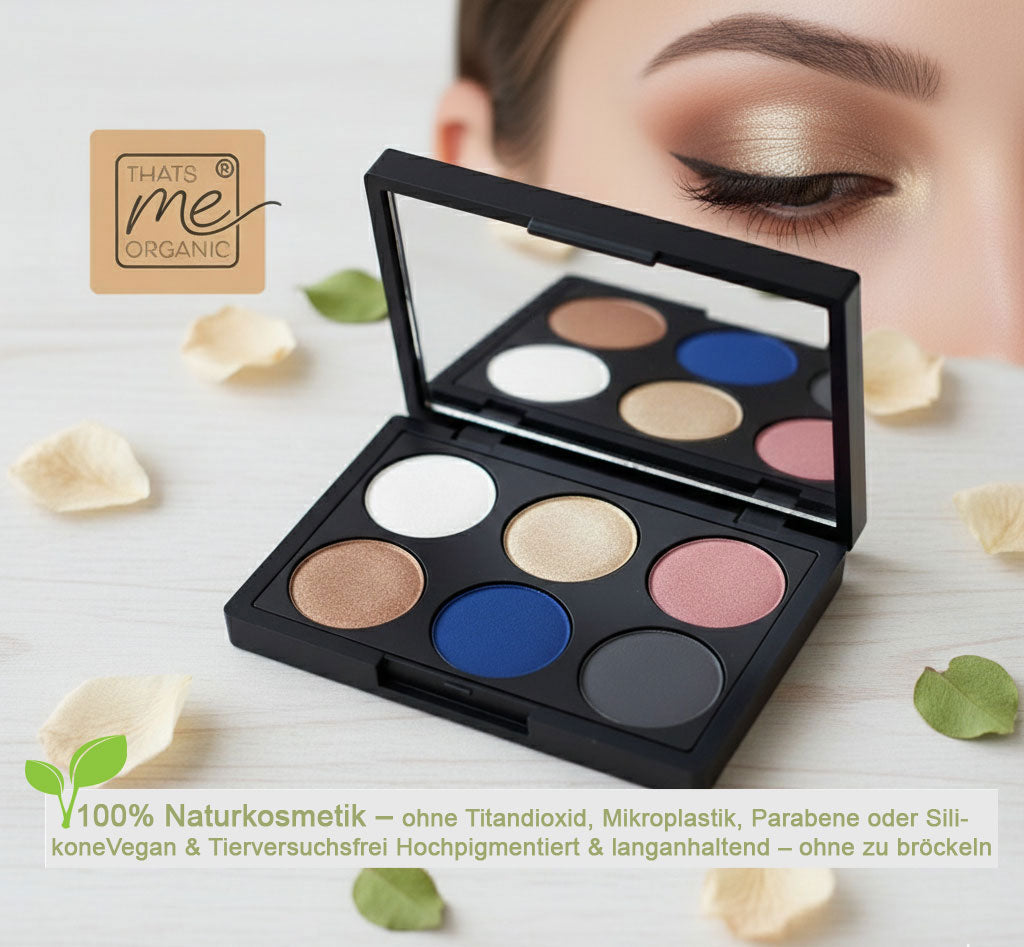 Eine offene schwarze Lidschattenpalette von "That's me organic" mit sechs kreisförmigen Farbtönen: Weiß, glänzendes Gold, mattes Blau, schimmerndes Bronze, Rosé und Anthrazit. Die Palette liegt auf einer hellen Holzoberfläche, umgeben von cremefarbenen Blütenblättern und grünen Blättern. Oben links ist das originale "That's me organic"-Logo. 100% Naturkosmetik – ohne Titandioxid, Mikroplastik, Parabene oder Silikone