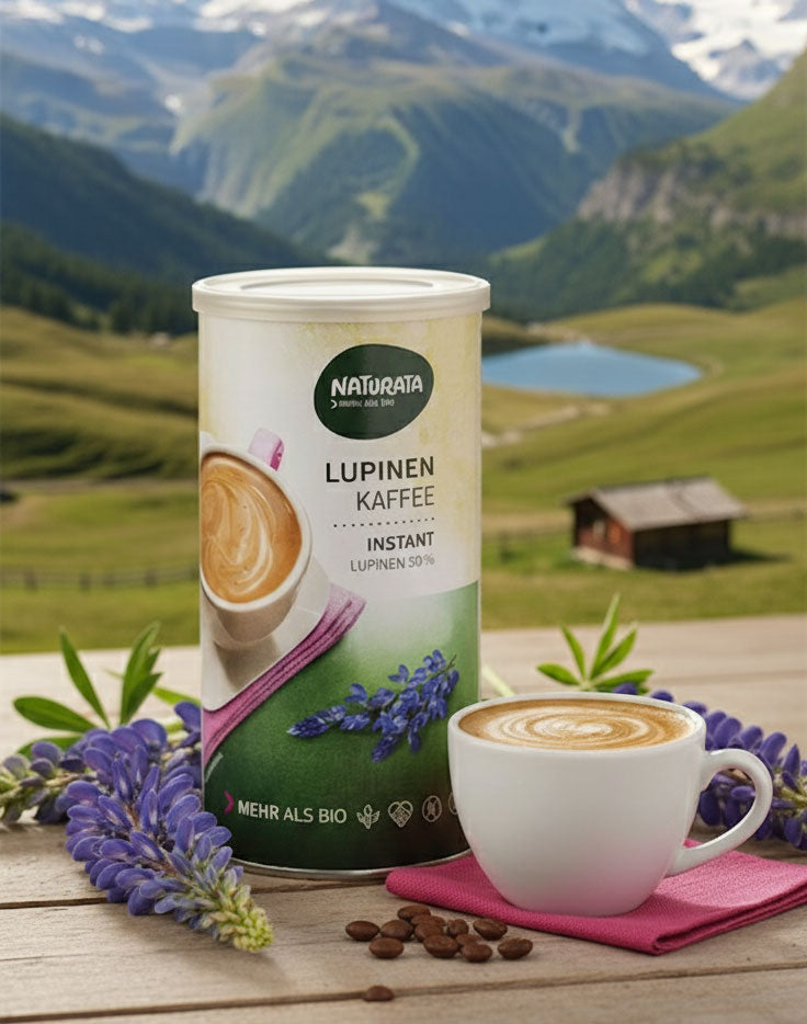 Eine Dose Naturata Lupinen Kaffee Instant steht auf einem rustikalen Holztisch. Links und rechts von der Dose sind frische, lilafarbene Lupinenblüten arrangiert. Vor der Dose steht eine weiße Tasse mit einem liebevoll zubereiteten Kaffee Latte, der auf einem pinken Tuch platziert ist. Vereinzelt liegen Kaffeebohnen auf dem Tisch. Im Hintergrund erheben sich majestätische, schneebedeckte Schweizer Berge, darunter das Matterhorn, sowie grüne Almen und ein kleiner See.