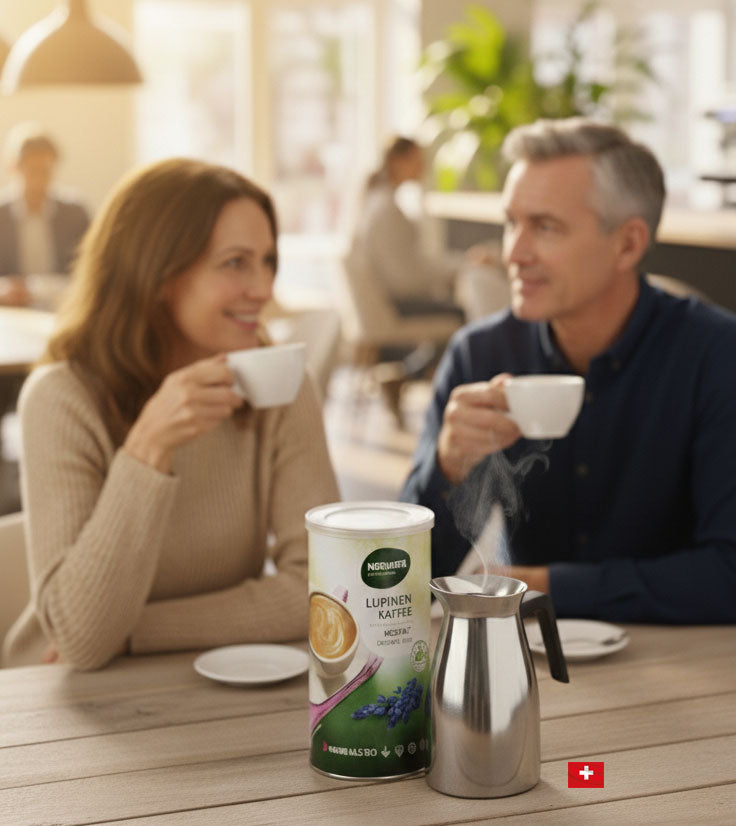 Auf einem rustikalen Holztisch steht eine Dose Naturata Lupinen Kaffee Instant neben einer dampfenden, silbernen Kaffeekanne. Vereinzelt liegen Kaffeebohnen auf dem Tisch. Im unscharfen Hintergrund sind ein Paar mittleren Alters zu sehen, das gemütlich Kaffee trinkt und sich unterhält, während sie an einem Tisch in einem hellen Café oder einer modernen Einrichtung sitzen. Die Szene ist von warmer, natürlicher Lichteinstrahlung durchdrungen.