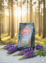 Eine dunkelgraue Packung 'lupino Kaffee aus Wertheimer Süßlupinen' steht auf einer hellen Oberfläche. Die Packung hat einen roten Kreis mit dem Text 'lupino' und 'immer und ewig' sowie einen blauen Streifen mit 'kein normaler kaffee'. Vor der Packung und zu den Seiten liegen mehrere Zweige mit violetten Lupinenblüten. Im unscharfen Hintergrund sind die Stämme eines Waldes bei Sonnenlicht zu sehen, wobei Sonnenstrahlen und leichte Partikel in der Luft schweben.