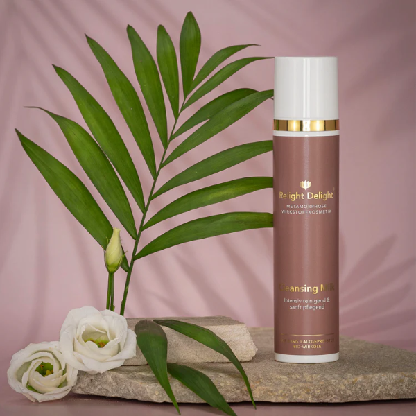 Edler Pumpspender in Altrosa mit goldener und weißer Aufschrift: Metamorphose Wirkstoffosmetik - Cleansing Milk 100ml Transformation die man sieht und fühlt - Relight Delight Auf Basis Kaltgepresster Bio-Wirköle. Daneben weiße Rosenköpfe, ein Palmblatt. Alle Produkte stehen auf einem hellen, flachen Stein.