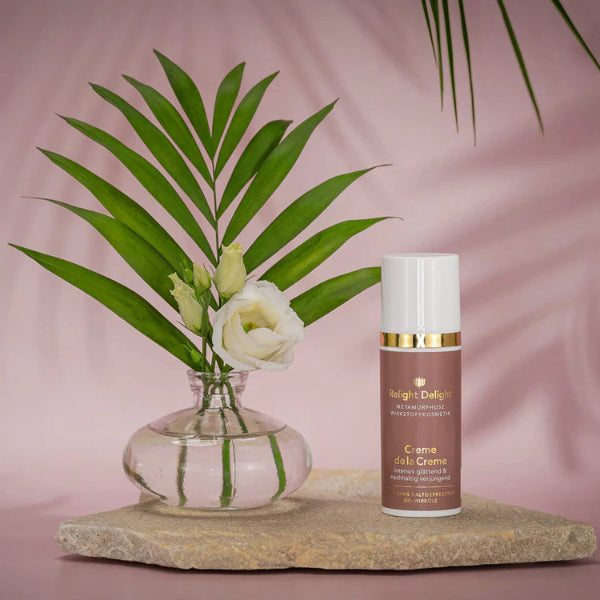 Edle Tube in Altrosa und daneben eine kleine Vase mit weißen Rosen. Auf der Tube in goldener Aufschrift: Metamorphose - Creme de la Creme - unbeduftet, Relight Delight