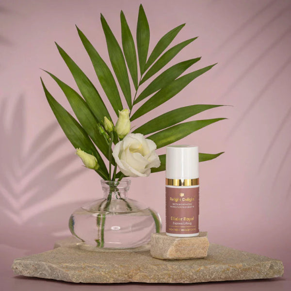 Edle Tube auf flachem, hellen Stein stehend des Metamorphose Elixier Royal Express Lifting  - 15ml  von Relight Delight, High Tech Wirkstoffe in Altrosa. Die Aufschrift ist in Gold und Weiß. Links daneben  eine edle Vase mit weißen Rosen