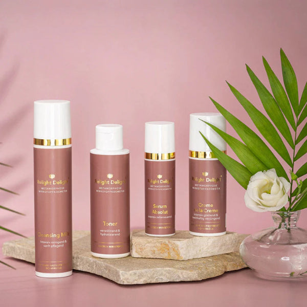 Edles Set des Metamorphose Gesichts-Set von Relight Delight bestehend aus Cleansink Milk, Toner, Serum Absolut und Creme de la Creme auf einem schnen hellen Stein gestellt, daneben eine Vase mit einer weißen Rose und einem Palmblatt