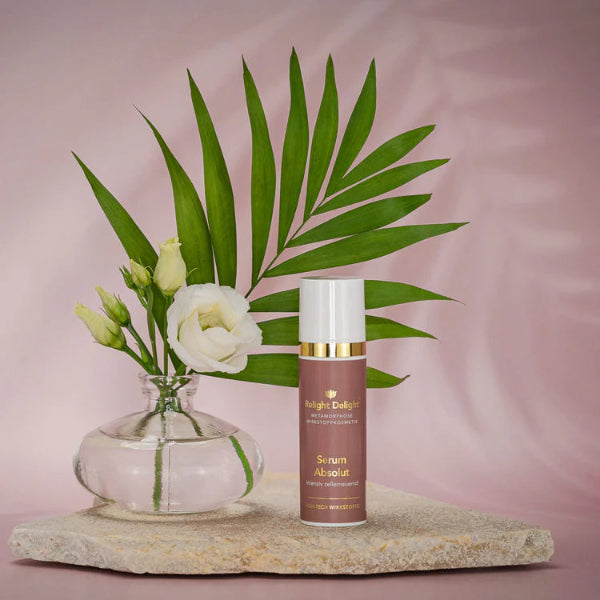 Tube in Altrosa mit goldener Aufschrift Metamorphose Wirkstoffkosmetik, Serum Absolut- frei von Parfüm 30ml -  Relight Delight steht qauf einem hellen flachen Stein. Links eine kleine Vase mit  weißen Rosen und ein Palmblatt. Der Hintergrund ist rosa gehalten