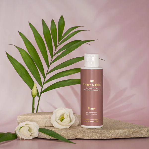 Schöne Tube in Altrosa mit goldener und weißer Schrift : Metamorphose Toner verwöhned und hydratisierend - Relight Delight - Gigh Tech Wirkstoffe steht auf hellem, flachen Stein. Daneben Rosenköpfe n weiß, ein Palmblatt und zartrosa Hintergrund