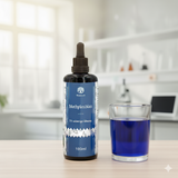 Eine Flasche Waldkraft Methylenblau 1% wässrige Lösung steht neben einem Glas mit blauer Flüssigkeit auf einem hellen Holztisch. Im unscharfen Hintergrund ist eine moderne Küchen- oder Laborumgebung mit einem Fenster und einigen Regalen zu sehen.