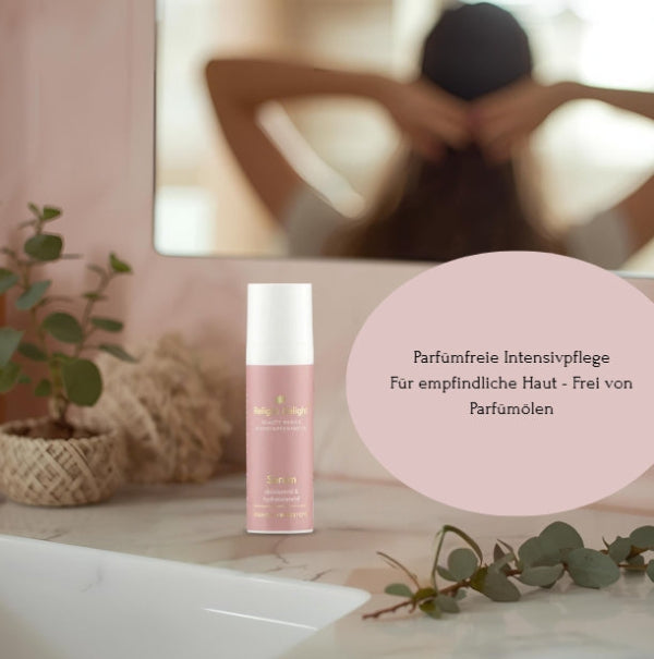 Im Vordergrund ist eine roséfarbene Tube Beauty Basics Serum auf einem weißen Marmorwaschtisch. Links davon befindet sich eine kleine Topfpflanze und rechts ein Eukalyptuszweig. Im Hintergrund, in einem Spiegel, ist unscharf eine Frau zu sehen, die ihr Haar im Nacken hält. Über dem Spiegel ist der Text „Parfümfreie Intensivpflege. Für empfindliche Haut - Frei von Parfümölen“ in einem ovalen rosafarbenen Feld zu sehen.