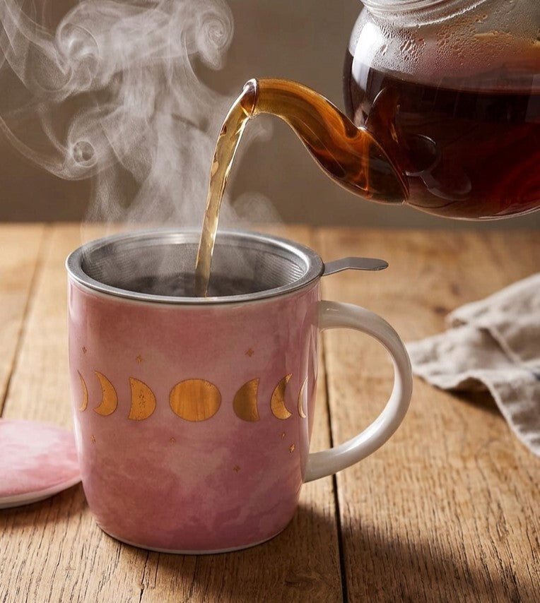 Heißer Tee wird aus einer Glaskanne in eine rosa Teetasse mit goldenen Mondphasen gegossen. Dampf steigt auf. Tasse  mit Deckel daneben.