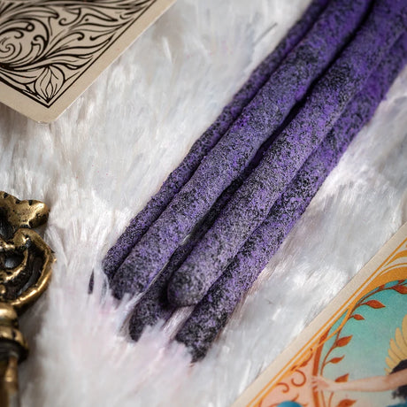 Nahaufnahme von mehreren lila Räucherstäbchen mit dicker, organischer Textur auf weißem Felluntergrund, umgeben von illustrierten Engelkarten.